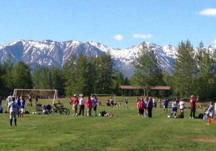 Snow capped mountains and blue sky behind green grass soccer field with players. 