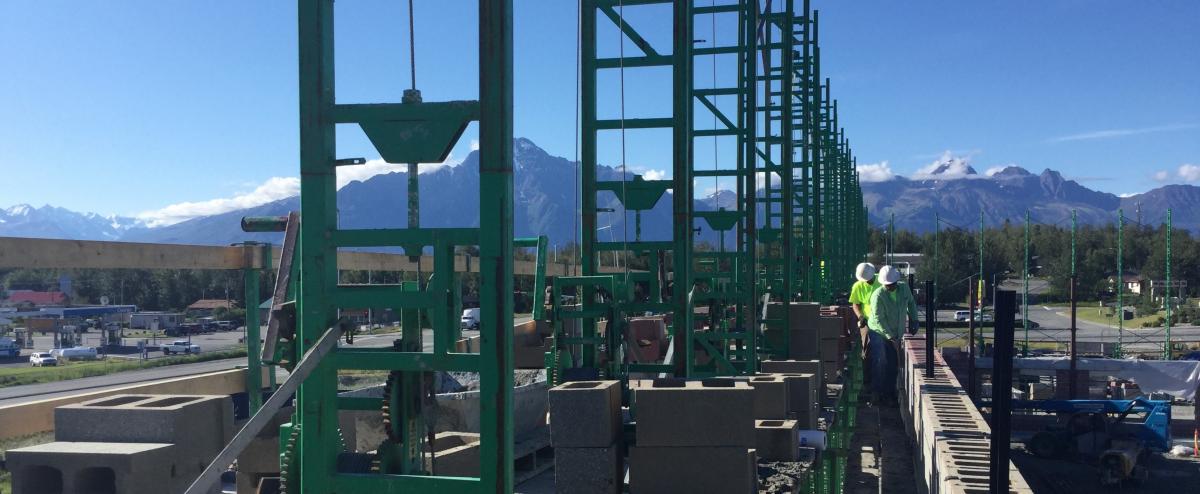 Cinderblock building under construction with green beams in the foreground and snow capped mountains in the background.
