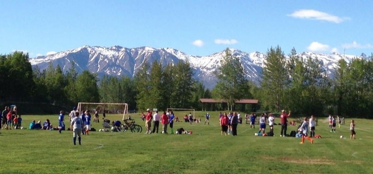 Snow capped mountains and blue sky behind green grass soccer fields with players. 